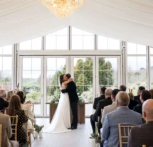 Couple sharing a first kiss at wedding ceremony in sunlit Fernhill House Hotel conservatory.