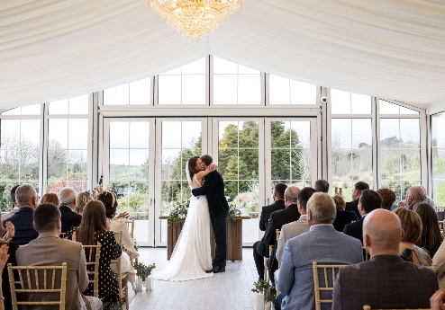A couple celebrates their wedding with a kiss in a bright, elegant conservatory, surrounded by family and friends.