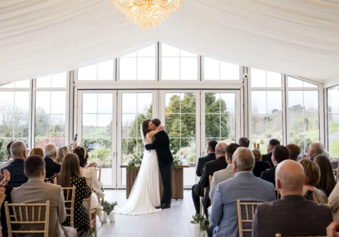 Couple sharing a first kiss at wedding ceremony in sunlit Fernhill House Hotel conservatory.