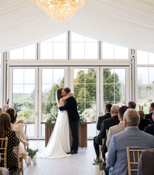A couple celebrates their wedding with a kiss in a bright, elegant conservatory, surrounded by family and friends.