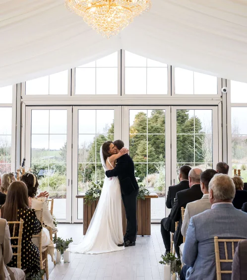 Couple sharing a first kiss at wedding ceremony in sunlit Fernhill House Hotel conservatory.
