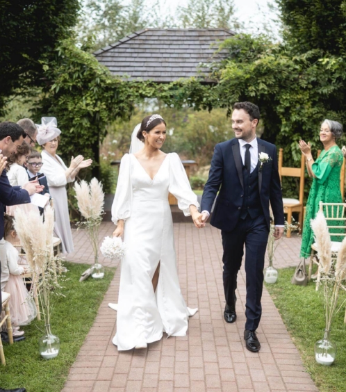A couple walks hand in hand down an outdoor aisle at Fernhill House Hotel, surrounded by joyful guests.
