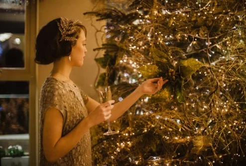 Woman in elegant dress holding champagne by a decorated Christmas tree.