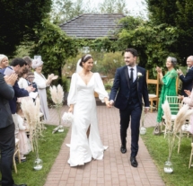 A joyful couple walks down an outdoor aisle at Fernhill House Hotel, surrounded by cheering family and friends.