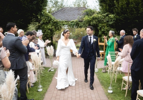 A joyful couple walks down an outdoor aisle at Fernhill House Hotel, surrounded by cheering family and friends.