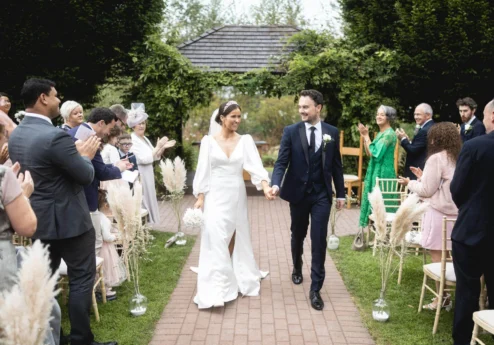 A joyful couple walks down an outdoor aisle at Fernhill House Hotel, surrounded by clapping family.