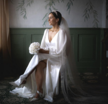 Bride in elegant gown sitting in a serene, softly lit room with floral wallpaper.