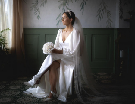 Bride in elegant gown sitting in a serene, softly lit room with floral wallpaper.