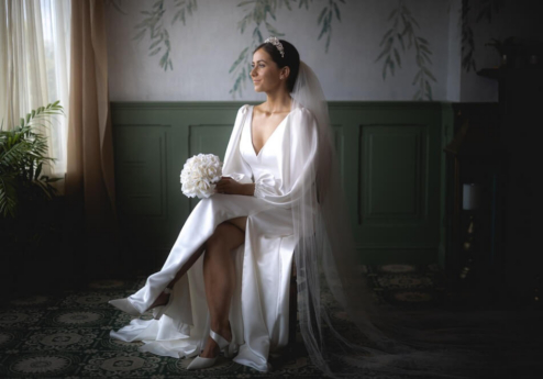 Bride in elegant gown sitting in a serene, softly lit room with floral wallpaper.