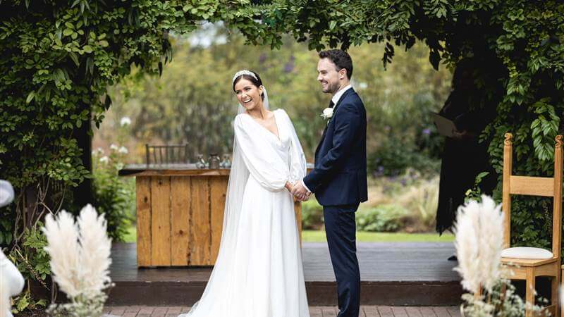 A couple stands smiling under a leafy archway at Fernhill House Hotel, ready for their wedding ceremony.