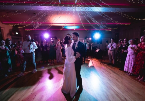 A couple dances at a wedding reception, surrounded by family under twinkling fairy lights.