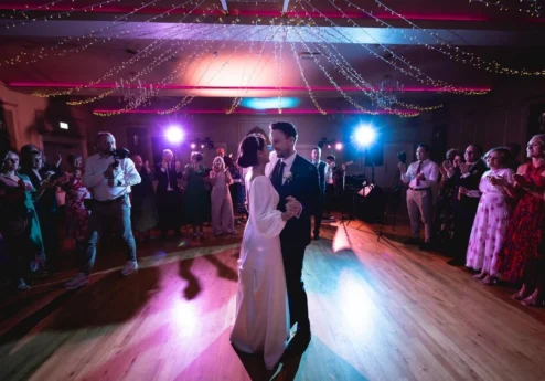 A couple enjoys a first dance in a warmly lit hotel ballroom, surrounded by family and friends.