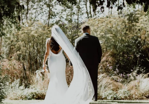 A couple in wedding attire strolls through lush gardens at Fernhill House Hotel.