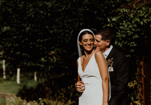 A couple embraces in a lush garden at Fernhill House Hotel, under bright sunlight.