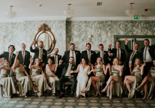 A joyous wedding party poses indoors, elegantly dressed in coordinated attire.