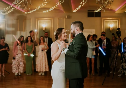 Couple dancing in an elegantly decorated ballroom with guests watching. Warm lighting and fairy lights.