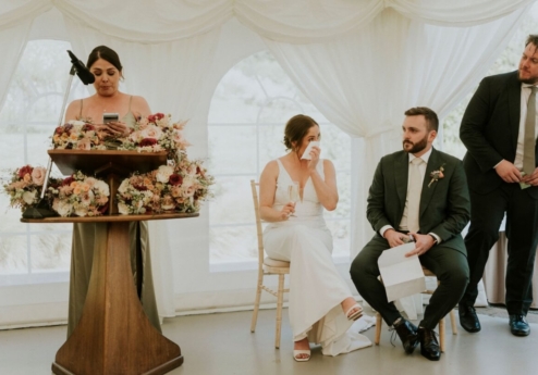 A couple seated in a decorated wedding marquee, listening to a speech.