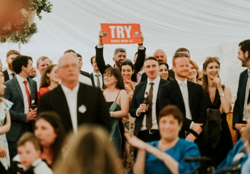 A joyful group of people in formal attire celebrating under a white marquee at Fernhill House Hotel. A man holds a red sign reading "TRY. BODIES NEVER LIE.