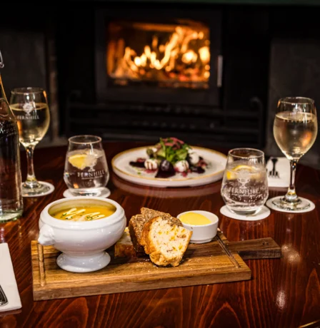 Cosy dining table with soup, salad, and drinks by a warm fireplace.
