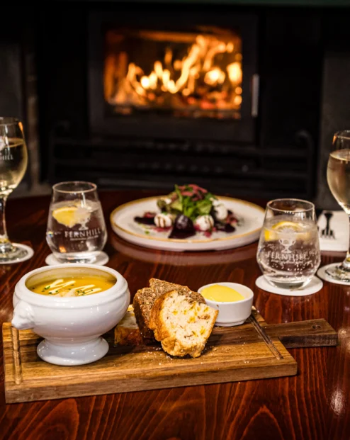 Cosy dining table with soup, salad, and drinks by a warm fireplace.