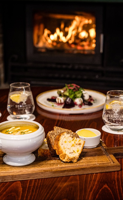 Cosy dining scene with soup, bread, and wine by a warm fireplace at Fernhill House Hotel.
