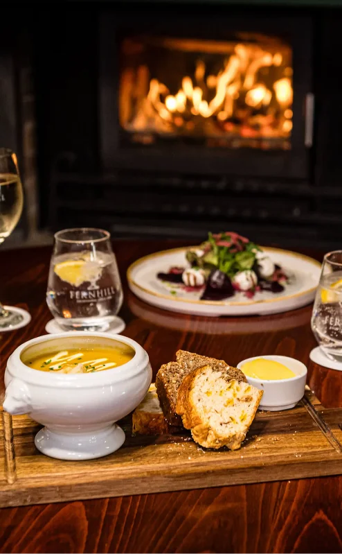 Cosy dining table with soup, salad, and drinks by a warm fireplace.