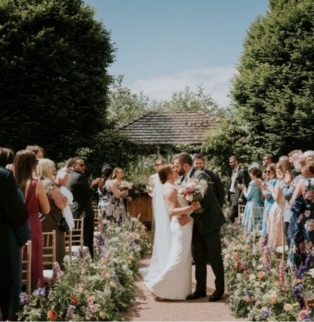 A joyful wedding couple kiss in a flower-lined garden, surrounded by applauding guests.