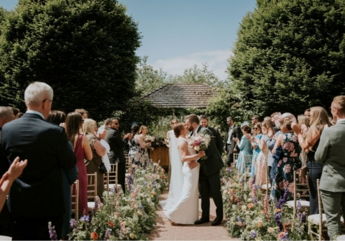 A joyful wedding couple kiss in a flower-lined garden, surrounded by applauding guests.