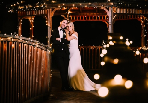 A smiling couple in wedding attire embrace under a gazebo decorated with fairy lights at night.