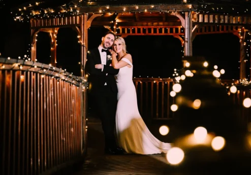 Romantic couple embraces on a fairy-lit wooden gazebo at Fernhill House Hotel.