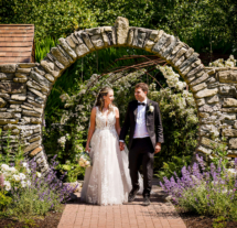 A couple in wedding attire stroll through a stone arch in a lush garden at Fernhill House.