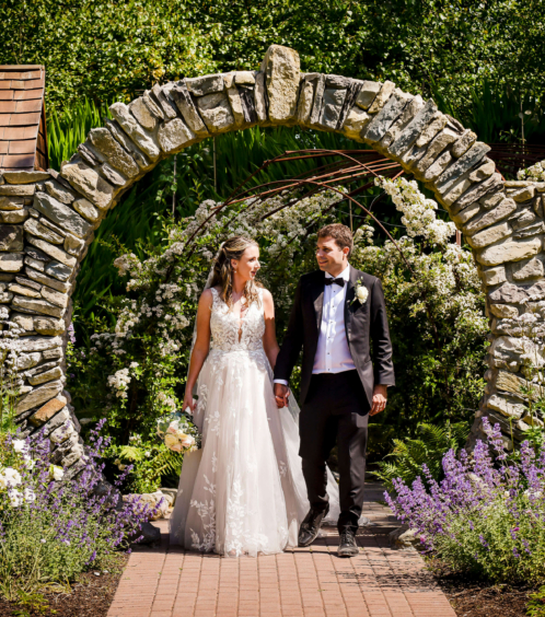 A couple in wedding attire stroll through a stone arch in a lush garden at Fernhill House.