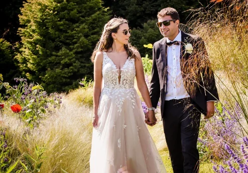 A couple in elegant attire walk hand in hand through a lush garden at Fernhill House Hotel.
