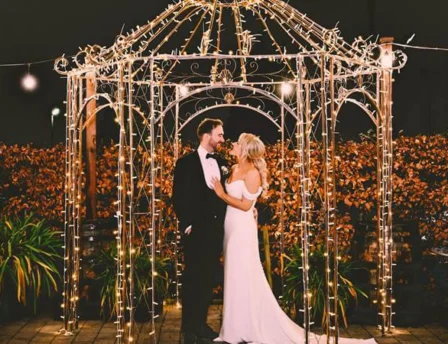 A couple embraces under a gazebo adorned with twinkling lights at night in a garden setting.