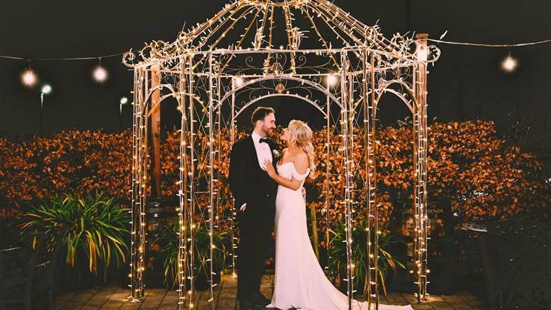 A couple embraces under a gazebo adorned with twinkling lights at night in a garden setting.
