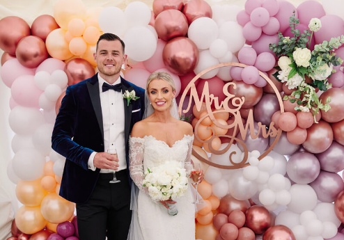 A couple smiling in formal attire in front of a balloon arch decorated with "Mr & Mrs" signage.