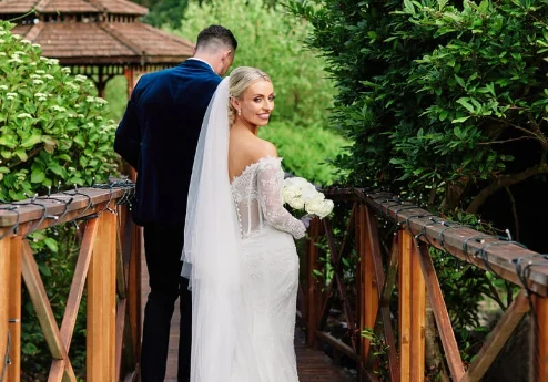 A couple in wedding attire walk over a wooden bridge surrounded by lush greenery.