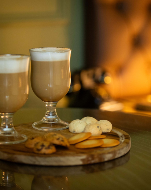 Cosy hotel room setting with two lattes and biscuits on a wooden tray.