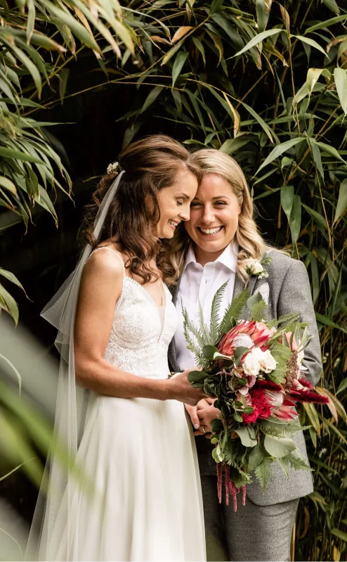 Couple embracing amid lush greenery at Fernhill House, holding a vibrant bouquet.