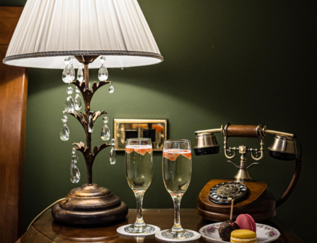 Cosy hotel room with vintage lamp, champagne, and macarons on a bedside table.