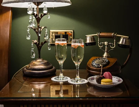 Elegant hotel room with lamp, vintage phone, champagne glasses, and macarons on a bedside table.