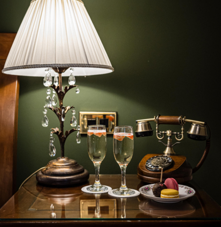 Cosy hotel room with vintage lamp, champagne, and macarons on a bedside table.