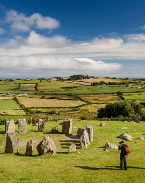 A couple explores an ancient stone circle amidst rolling green hills under a blue sky in the countryside.