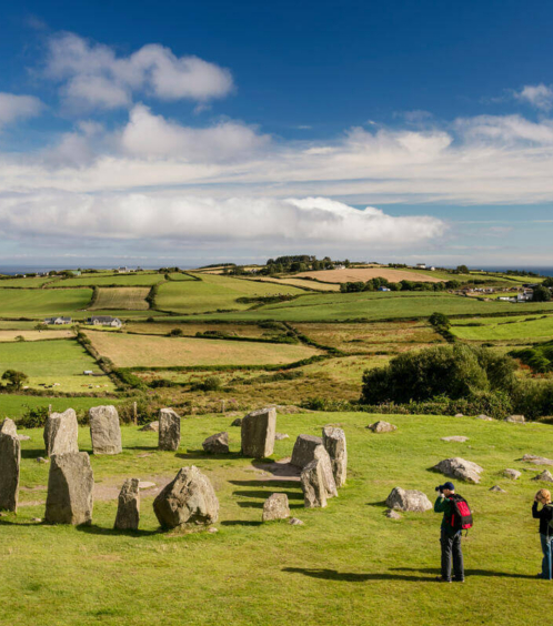 A couple explores an ancient stone circle amidst rolling green hills under a blue sky in the countryside.