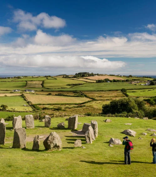 A couple exploring an ancient stone circle in a lush, green countryside under a blue sky.