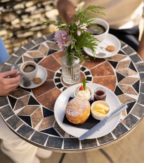 Couple enjoying coffee and pastries at a mosaic outdoor table with flowers.