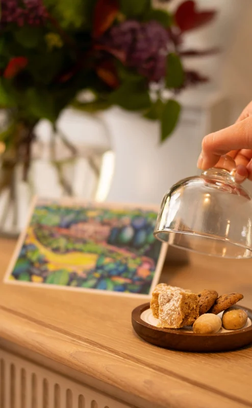 Hand lifting cloche from plate of biscuits on wooden table near flowers and scenic postcard.