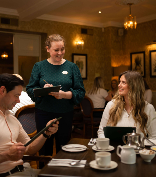 Friendly waiter serving a couple in a warm, inviting Fernhill House Hotel dining room.