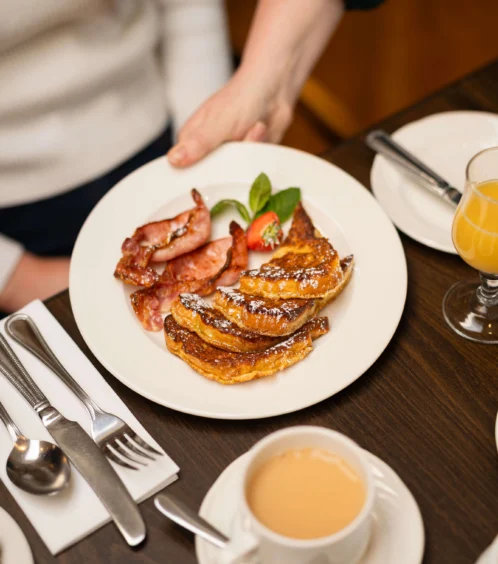 A person serves a plate of French toast and bacon with tea and juice at a cosy breakfast table.