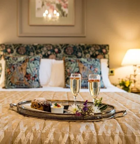 Elegant hotel room with a tray of desserts and champagne on the bed, soft lighting.